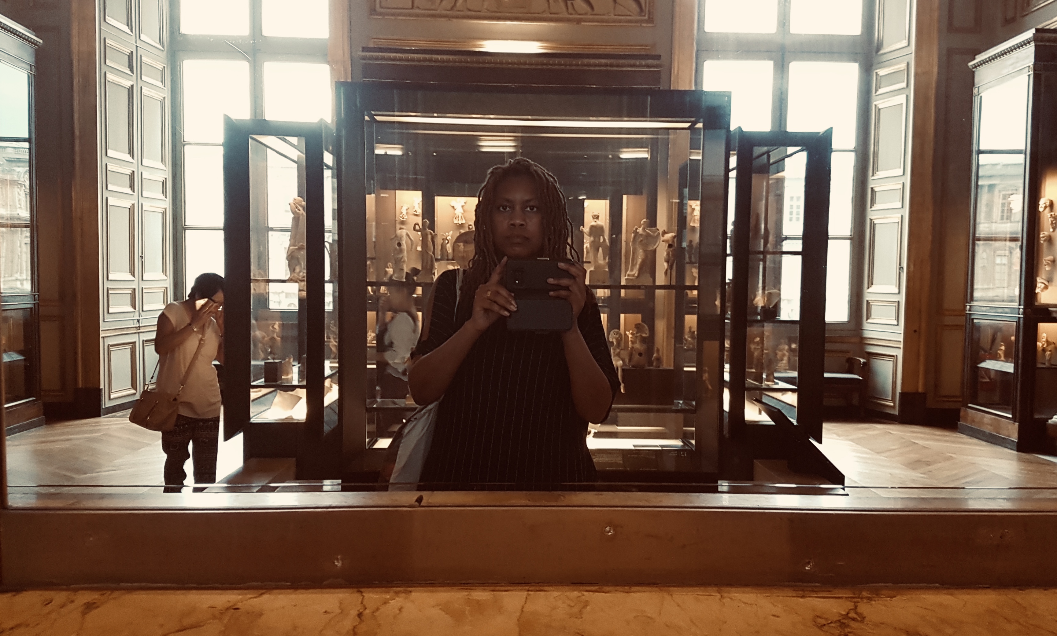 Mirror self-portrait in the Louvre's terracotta rooms: phone raised between the panes of a black vitrine full of small antique figures, a visitor rubbing her eyes at left
