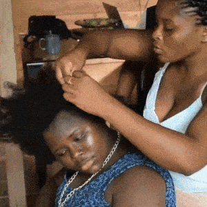 Protective styles: hands parting and braiding a seated girl's hair, her eyes patient and downcast, a mug and dark shelf behind