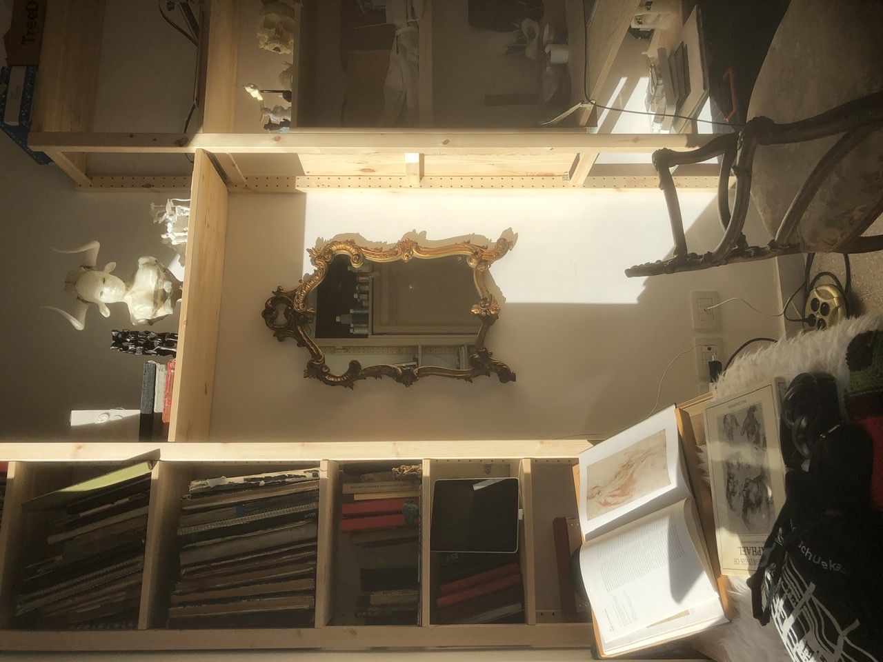 Roman studio in morning sun (photo on its side): gilt rococo mirror on a bare wall, pine shelving with white sculptures and skull, an open notebook of red-chalk drawing