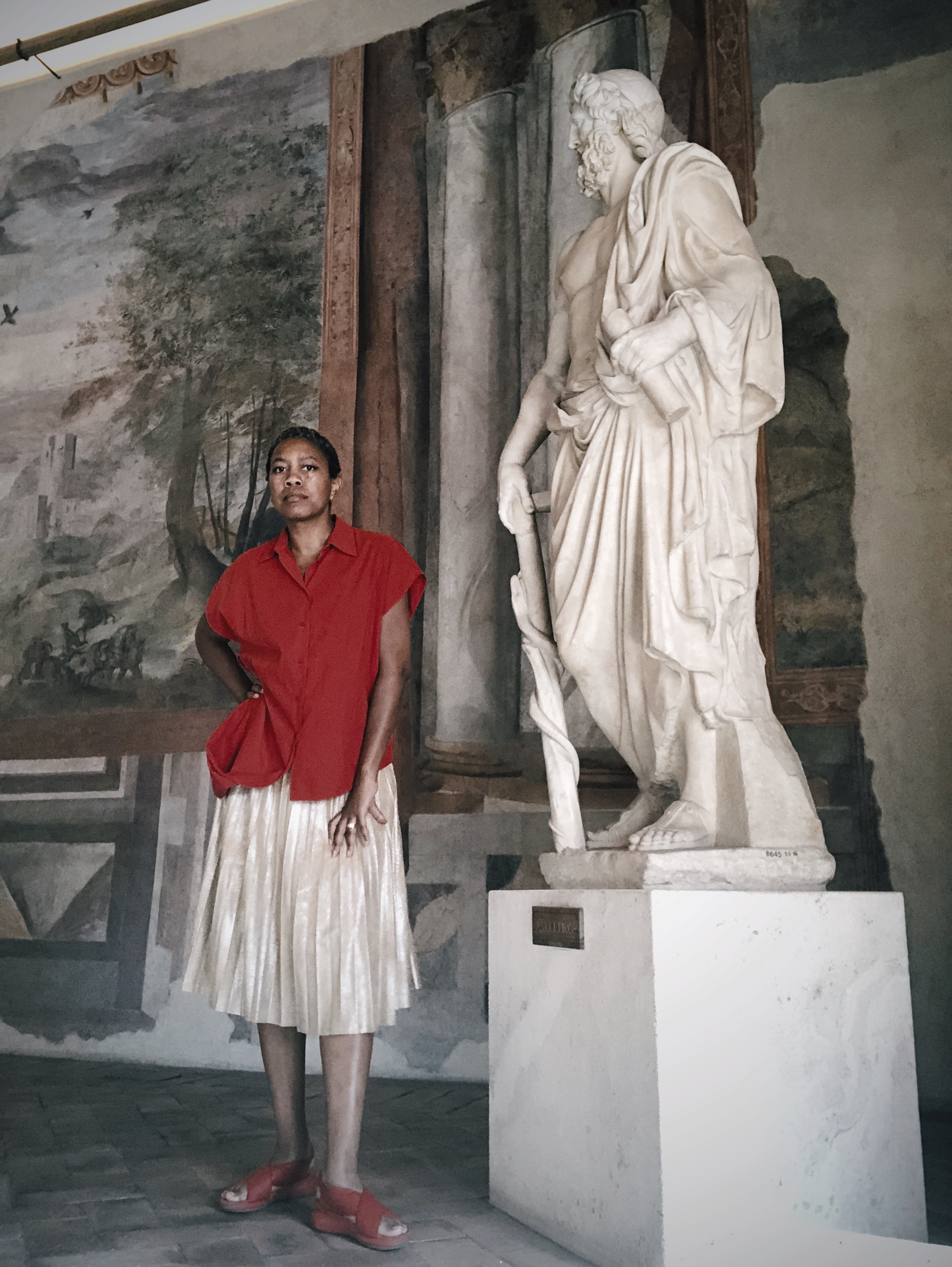 Standing hand-on-hip in red shirt and gold pleated skirt beside a marble Asklepios, in front of a frescoed landscape wall in Rome Standing hand-on-hip in red shirt and gold pleated skirt beside a marble Asklepios, in front of a frescoed landscape wall in Rome
