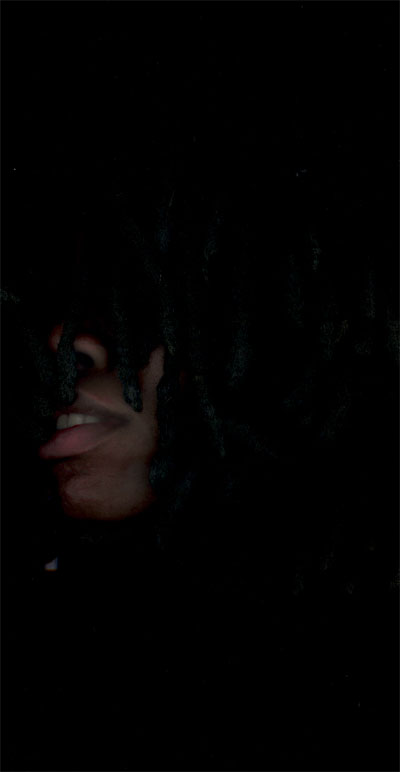 Portrait swallowed by darkness: only the tip of a nose, a bared smile and chin lit between black locs