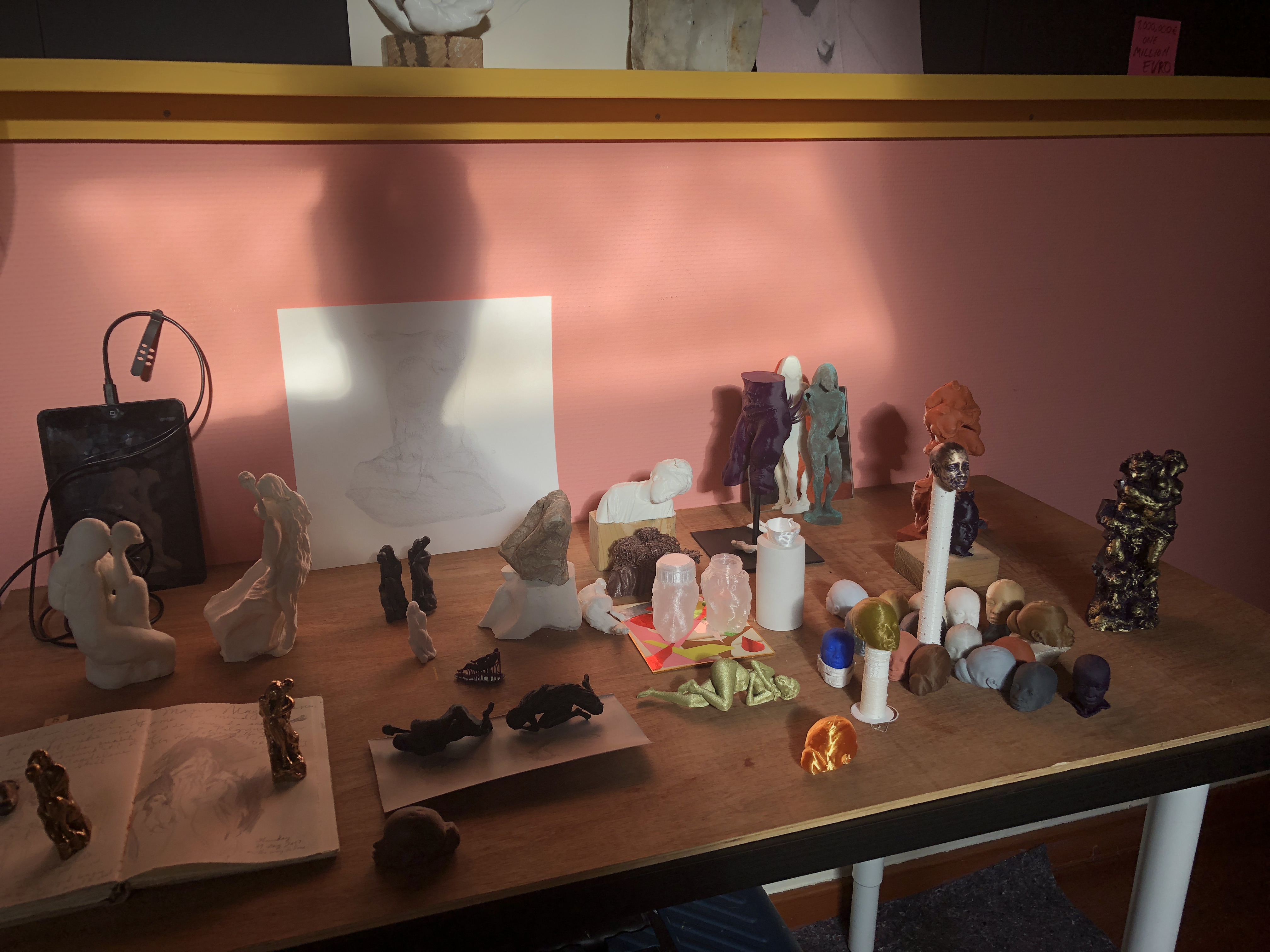 The print studio bench against pink walls: a crowd of small sculptures — printed heads in a dozen materials, black falling figures, gold twins, glass jars — with an iPad, a pencil study, and a sticky note pricing it all at ONE MILLION EURO