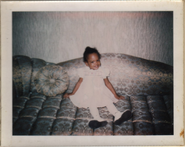 Polaroid from the seventies: a small girl in a white dress and tights grinning on a huge brocade sofa, feet not reaching the floor