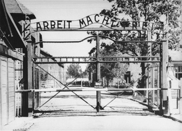 Black-and-white photograph of the Auschwitz gate, the wrought-iron ARBEIT MACHT FREI arch over the empty road between barracks