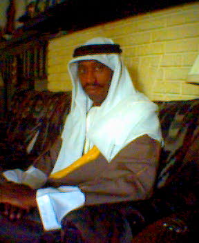 Barry back from Iraq: seated on the couch in a white keffiyeh and brown robes, photographed in warm low light