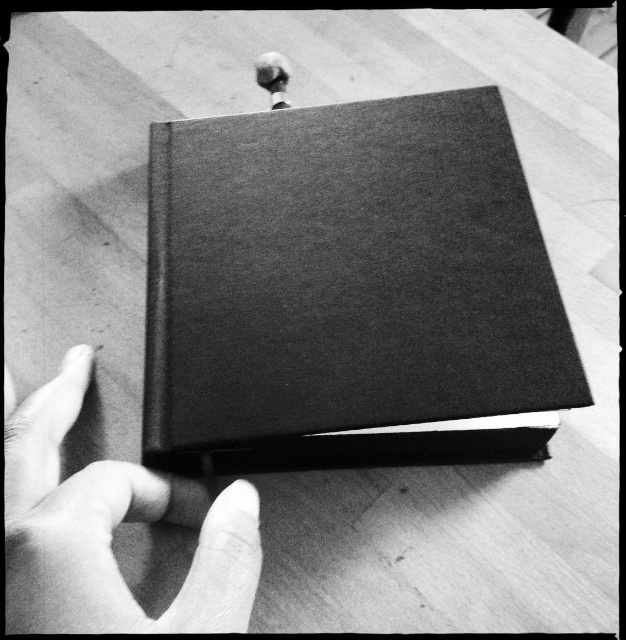Sketchbook 45 before its cover painting: a black-and-white photo of the plain black square book held at one corner, a pencil resting behind it