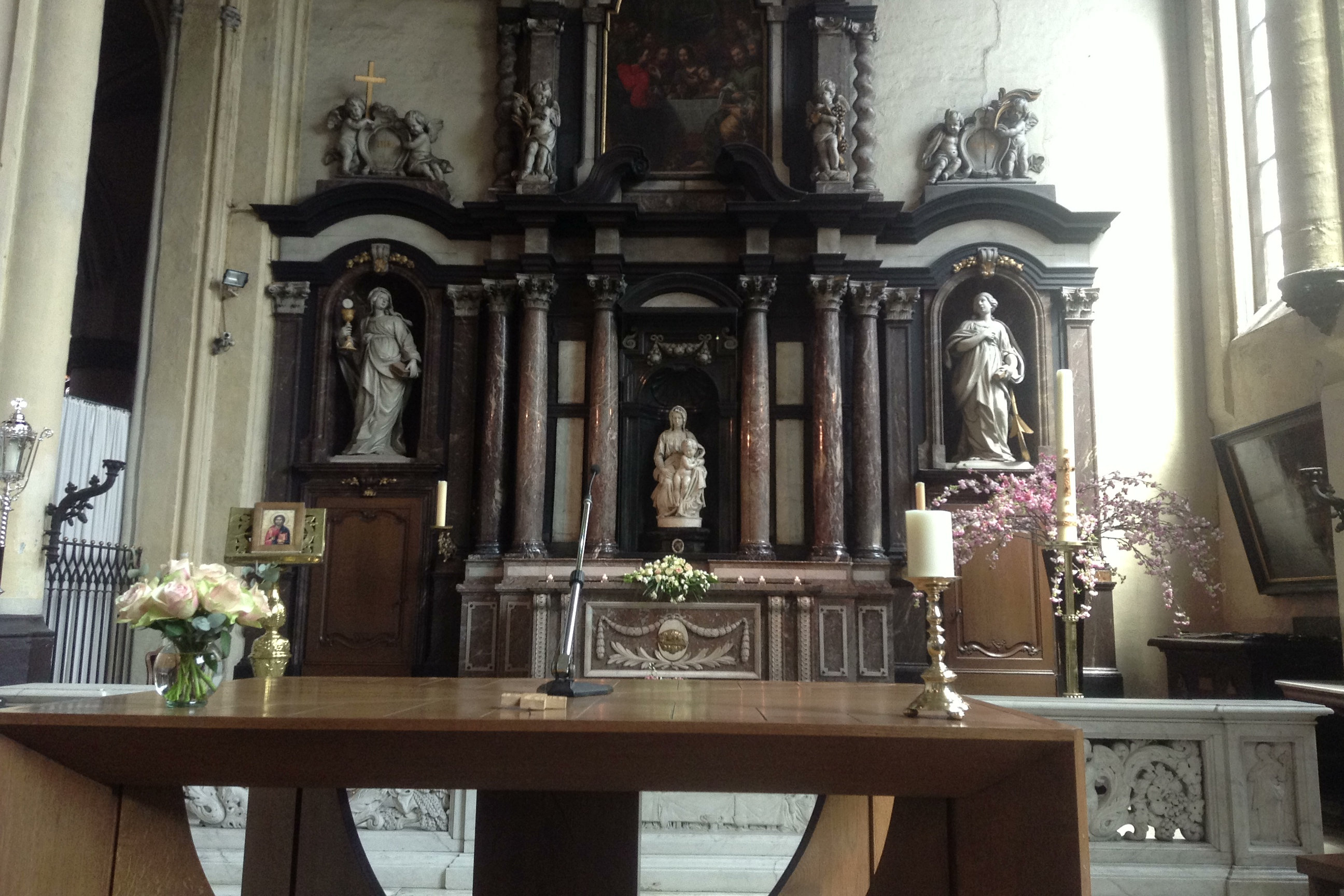 Michelangelo's Madonna of Bruges in her dark marble altar niche at the Church of Our Lady, flanked by saints, candles, roses and pink blossom branches Michelangelo's Madonna of Bruges in her dark marble altar niche at the Church of Our Lady, flanked by saints, candles, roses and pink blossom branches