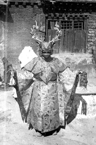Black-and-white photograph of a Tibetan cham dancer in an antlered deer mask and heavy brocade robes, arms spread outside a monastery door