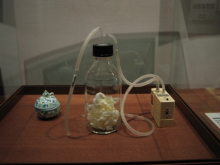 Museum vitrine with a device to measure the scent of a flower: white blossoms sealed in a glass bottle, tubing running to a small wooden pump box beside a porcelain jar
