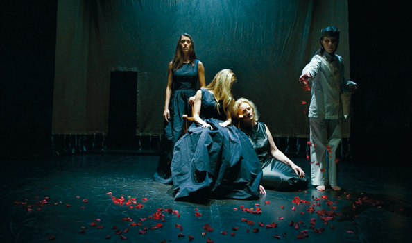 Stage photo from a Dido and Aeneas production: three women entwined around a spreading dark-blue gown while a figure in white scatters red rose petals across the black floor