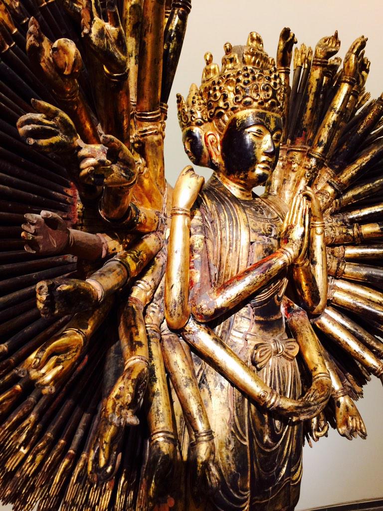 Gilded thousand-armed statue: a crowned deity with hands joined in prayer, a great fan of golden arms and open palms radiating behind the dark serene face