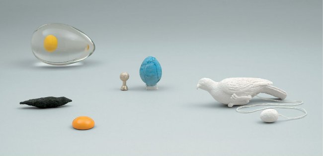 Still life of small art objects on grey: a glass egg with suspended yolk, a black bird, an orange yolk, a turquoise egg, a tiny figure and a white bird pull-toy on a string