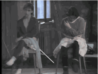 Grainy black-and-white video still from Rosas' Elena's Aria: two women seated on wooden chairs against a bare wall, one crossing her legs, one clutching her shoulder