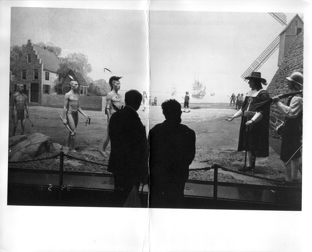Black-and-white photograph of two visitors silhouetted before a museum diorama of New Amsterdam: Lenape men with clubs face Dutch colonists by a windmill and stone house