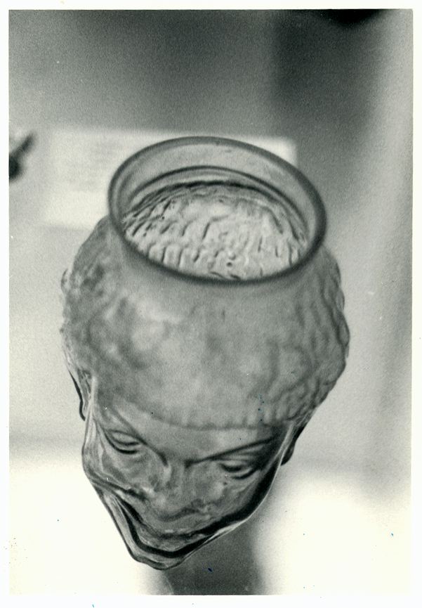 Black-and-white museum photograph of an ancient mold-blown glass flask shaped as a human head, seen from above so the open rim gapes where the crown should be
