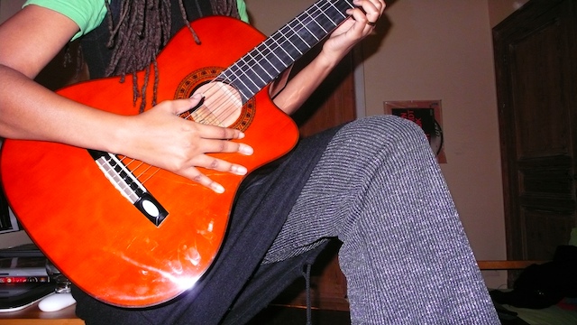 Auriea seated with an orange classical guitar, dreadlocks falling over a green tee, right hand spread across the strings