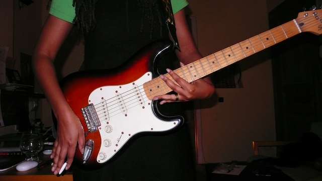 Auriea standing with a sunburst electric guitar in a dark room, one hand on the maple neck, a cigarette between the fingers of the other