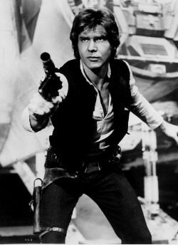 Black-and-white still of Harrison Ford as Han Solo, crouched with blaster drawn in front of the Millennium Falcon