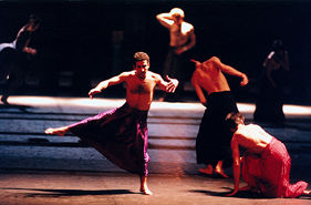 Stage photo from Wim Vandekeybus's In Spite of Wishing and Wanting: bare-chested men in fuchsia skirts, one balancing in a deep arabesque, others crouched and turning