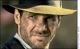 Harrison Ford as Indiana Jones in close-up under the brim of his fedora, face smudged with dirt, smiling sidelong