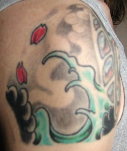 Shoulder tattoo in progress: green Japanese waves curling around dark smoke-grey clouds, two red petals falling, partly outlined panels near the shoulder blade