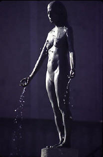 Kiki Smith's bronze fountain figure: a standing nude woman on a pedestal, water trickling from her open hands, photographed against violet light