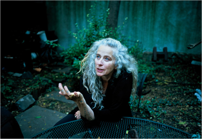Portrait of sculptor Kiki Smith in her overgrown back garden, grey curls loose, tattooed hand raised mid-thought