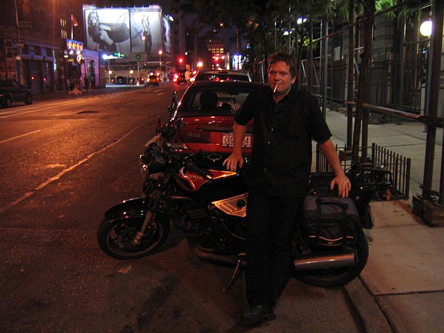 Night photo of Kit Halsted, cigarette in mouth, leaning on a loaded Triumph motorcycle at a Manhattan curb, lit billboards down the street