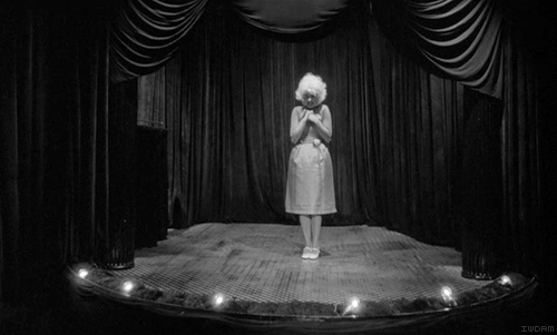 Animation frames from Eraserhead: the pale-cheeked Lady in the Radiator standing with clasped hands on her small lit stage between dark curtains
