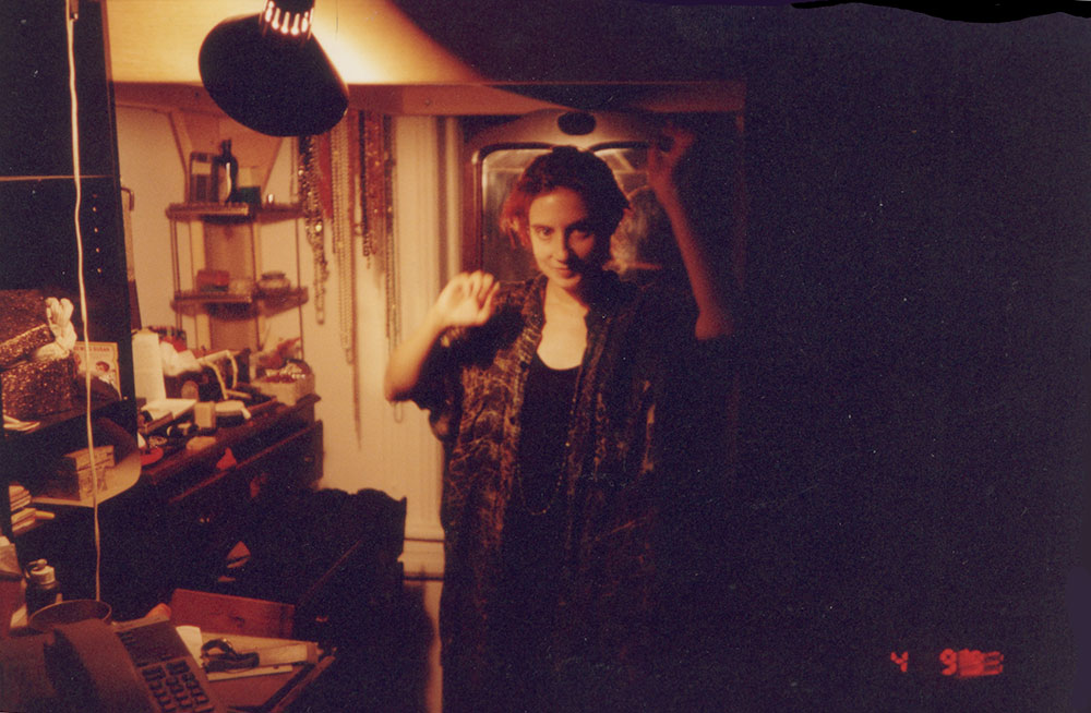Snapshot dated April 1993: a young woman in a patterned vest smiles under a desk lamp in a cluttered dorm room, necklaces hanging on the wall behind Snapshot dated April 1993: a young woman in a patterned vest smiles under a desk lamp in a cluttered dorm room, necklaces hanging on the wall behind