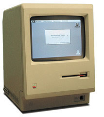 The original beige Apple Macintosh: built-in monochrome screen showing a welcome dialog, floppy slot below, keyboard cable in front