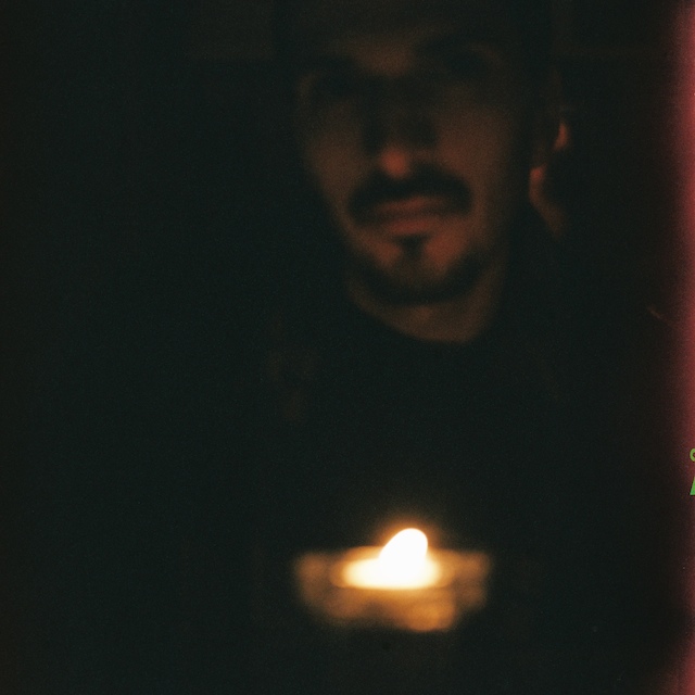Near-black photograph of a bearded man's face barely lit from below by a single tealight flame