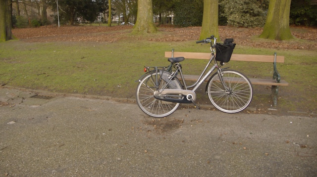 A grey Dutch bicycle with a black basket leaning against a park bench in a winter park, bare trees and drifts of fallen leaves behind