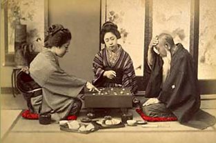 Hand-tinted Meiji-era photograph of two women and an older man gathered around a game of Go on a low board, tea things beside them