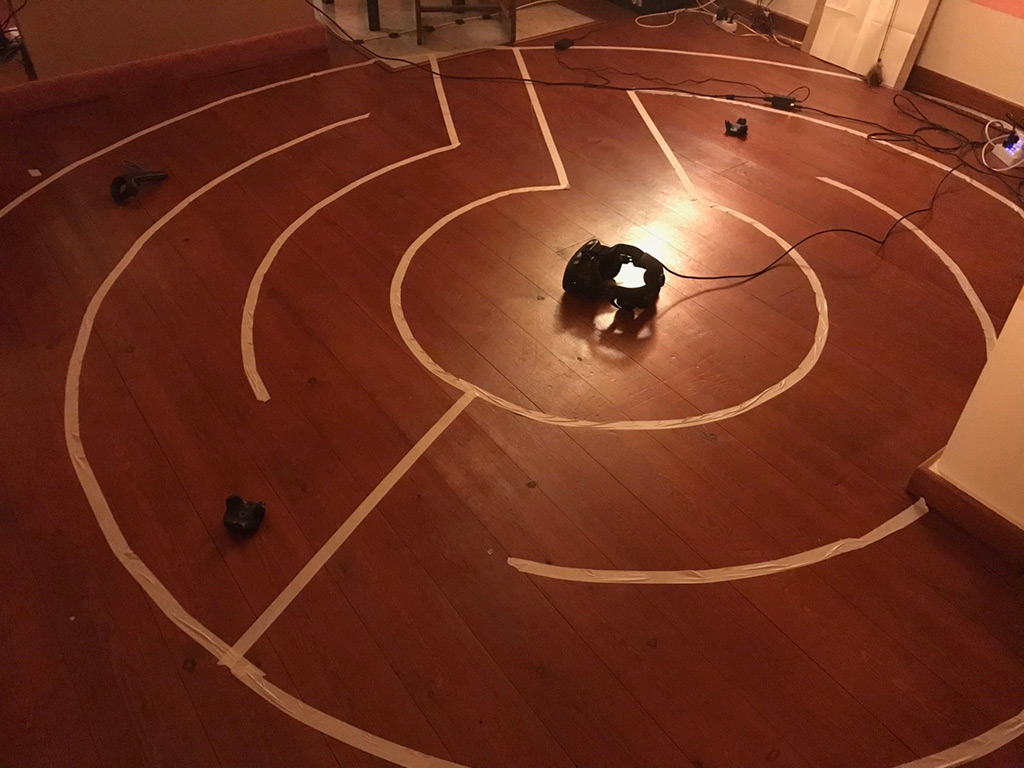 The MinoRinth play space in the studio: a spiral labyrinth taped in white on a wooden floor, an HTC Vive headset resting at its lit center, controllers at the edges The MinoRinth play space in the studio: a spiral labyrinth taped in white on a wooden floor, an HTC Vive headset resting at its lit center, controllers at the edges