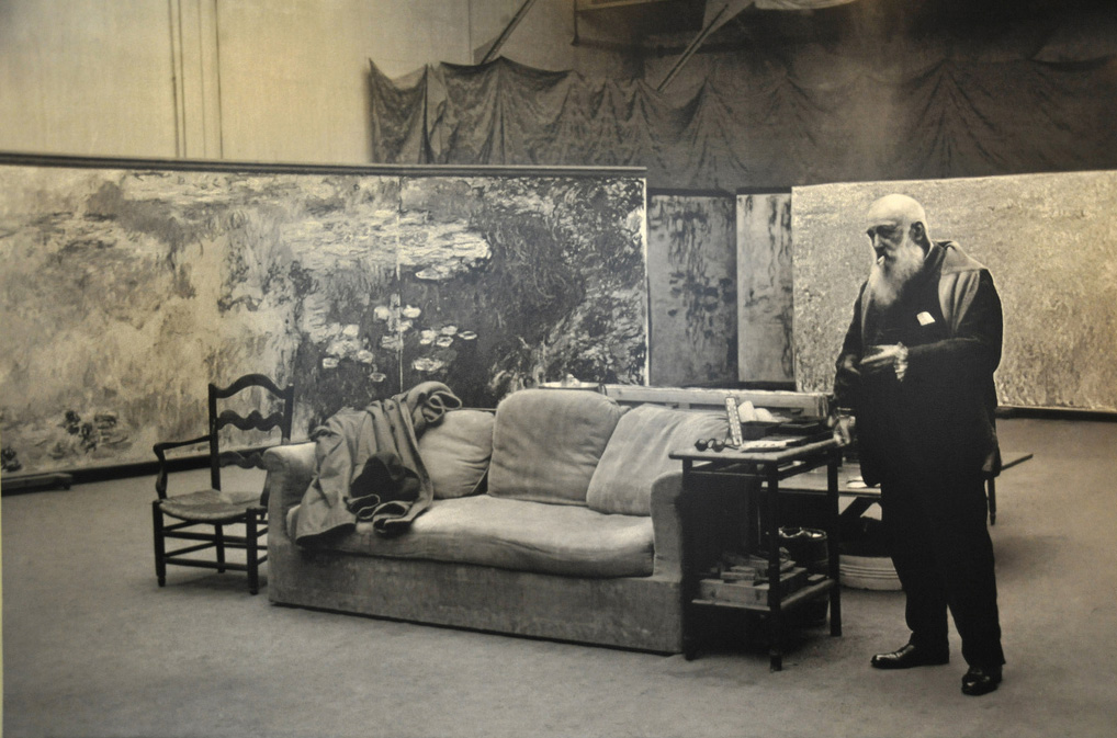 Black-and-white photograph of Claude Monet standing in his Giverny studio beside a sofa, surrounded by the huge water-lily panels