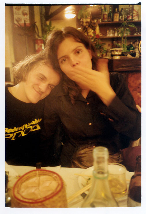 Olia Lialina and Dragan Espenschied leaning together at a restaurant table, she blowing a kiss past a bottle, a wheel of cheese in the foreground