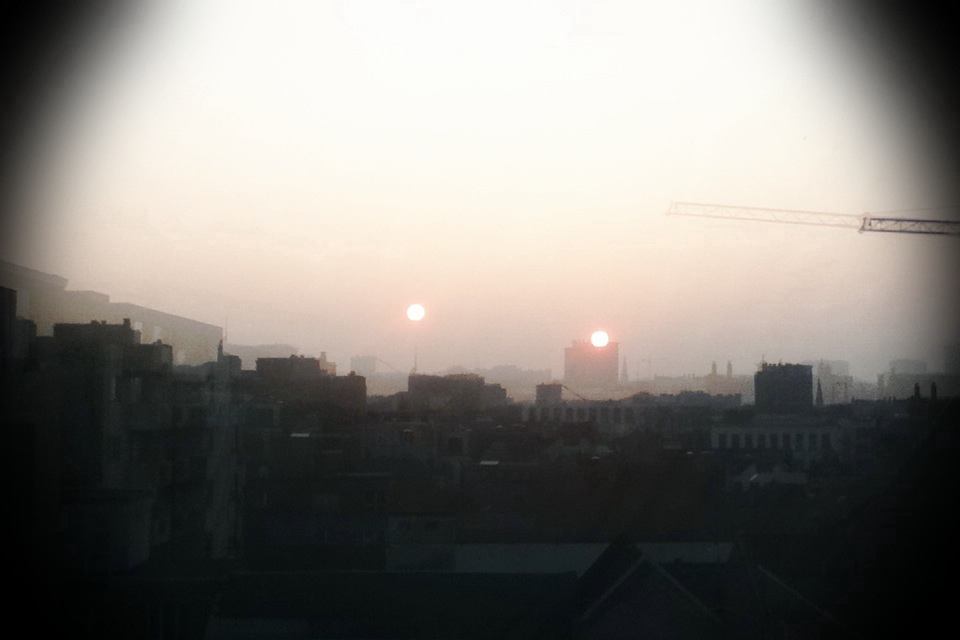 Hazy sunset over Ghent rooftops seen from the Pollepelstraat window: two pale suns — one real, one reflected — hang over silhouetted buildings and a crane