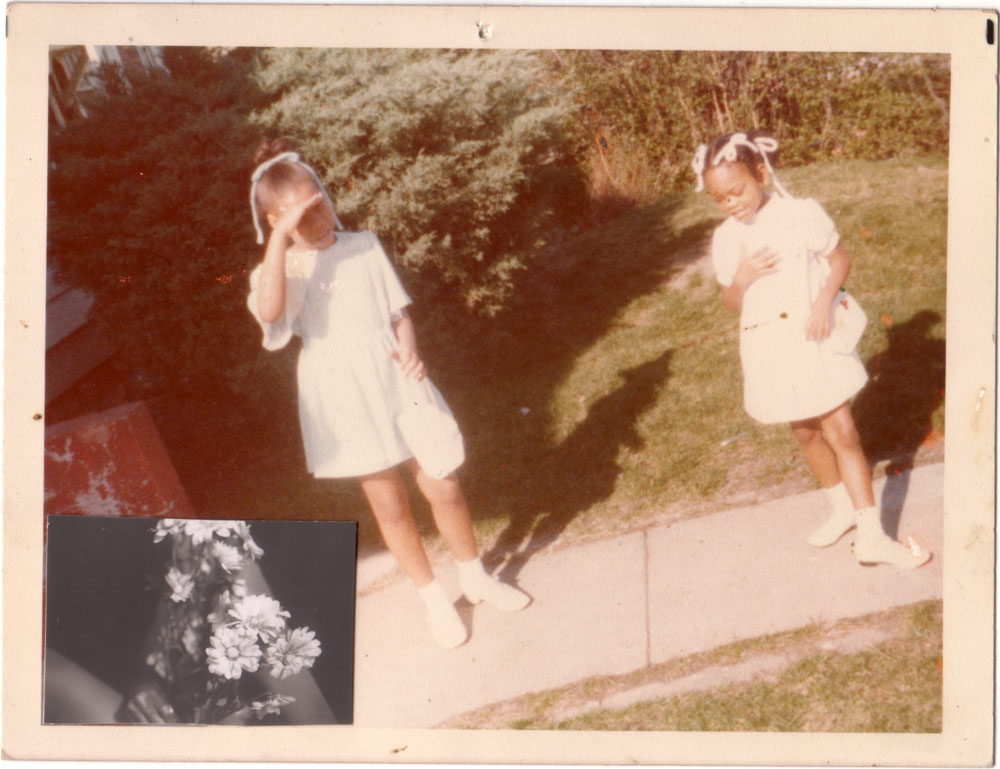 Faded 1970s snapshot of two small girls in white Easter dresses and hair ribbons on a sunny sidewalk, a black-and-white photo of daisies tucked into the corner