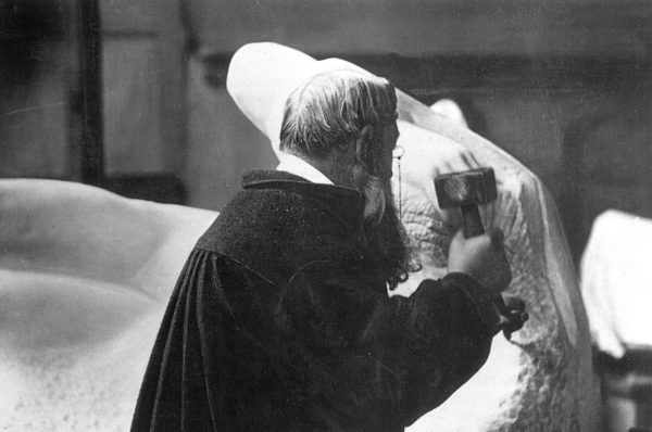 Black-and-white photograph of Auguste Rodin, bearded and in a dark smock, raising a mallet against a huge white marble block
