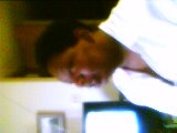 Tiny blurry webcam frame of Shari in a white shirt, head bowed over a desk in daylight