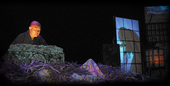 Theatrical scene lit against black: a bald monk figure peers over a carved stone chest across a floor of purple tangles toward a shoji screen with a shadowed silhouette Theatrical scene lit against black: a bald monk figure peers over a carved stone chest across a floor of purple tangles toward a shoji screen with a shadowed silhouette