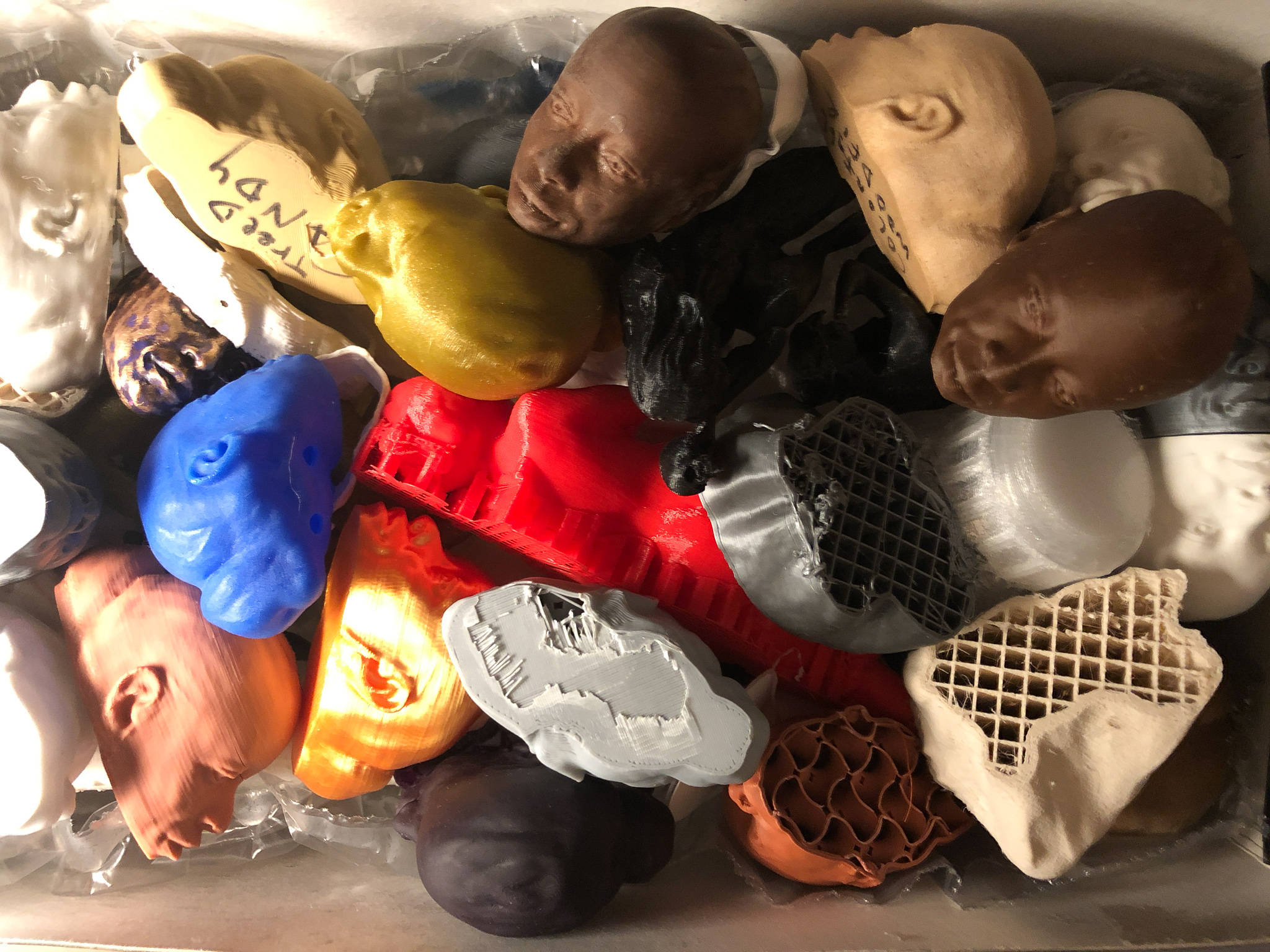 A box crammed with 3D-print tests: faces and heads in chocolate brown, blue, gold, copper, and bone, red architectural fragments, and gridded lattice scraps A box crammed with 3D-print tests: faces and heads in chocolate brown, blue, gold, copper, and bone, red architectural fragments, and gridded lattice scraps