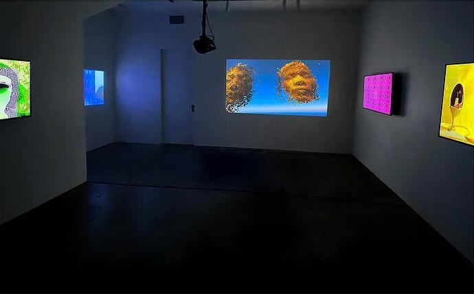 The Archive to Come at Telematic gallery: a dark room where a projection of two golden particle faces hovers on blue between bright screens on the walls
