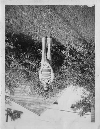 Black-and-white snapshot printed upside down: a figure in a striped top stands on a lawn before shrubs, inverted like the tarot Hanged Man