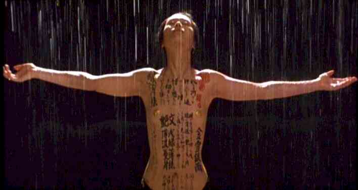Still from Peter Greenaway's The Pillow Book: a body covered in black calligraphy, arms spread wide under falling rain against darkness