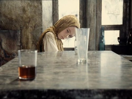 Still from Tarkovsky's Stalker: the Stalker's daughter in a gold headscarf rests her head beside two glasses on a stone table lit by a window