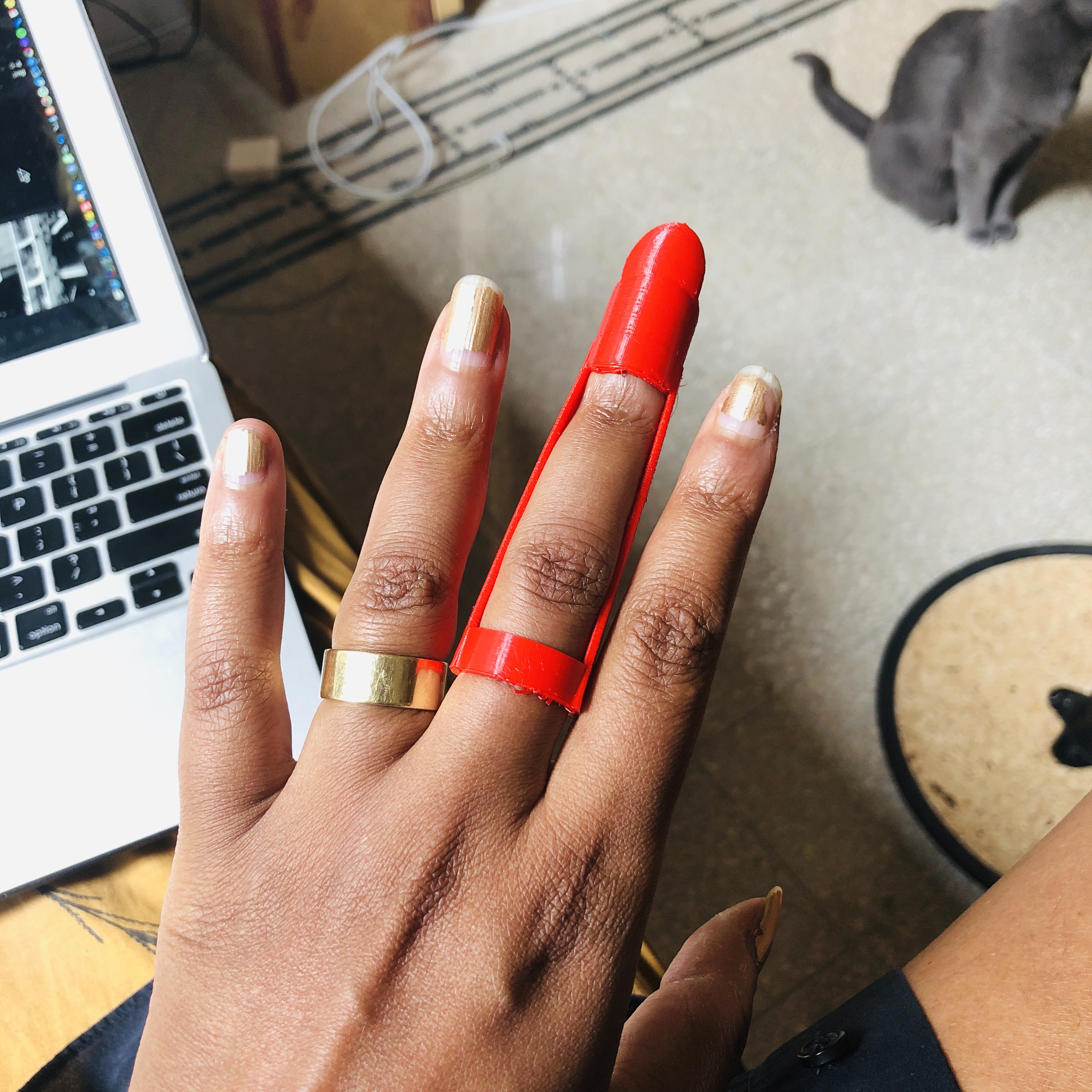 A red 3d-printed extension worn over the middle finger of a brown hand with gold-painted nails and a gold ring; laptop keyboard and grey cat beyond