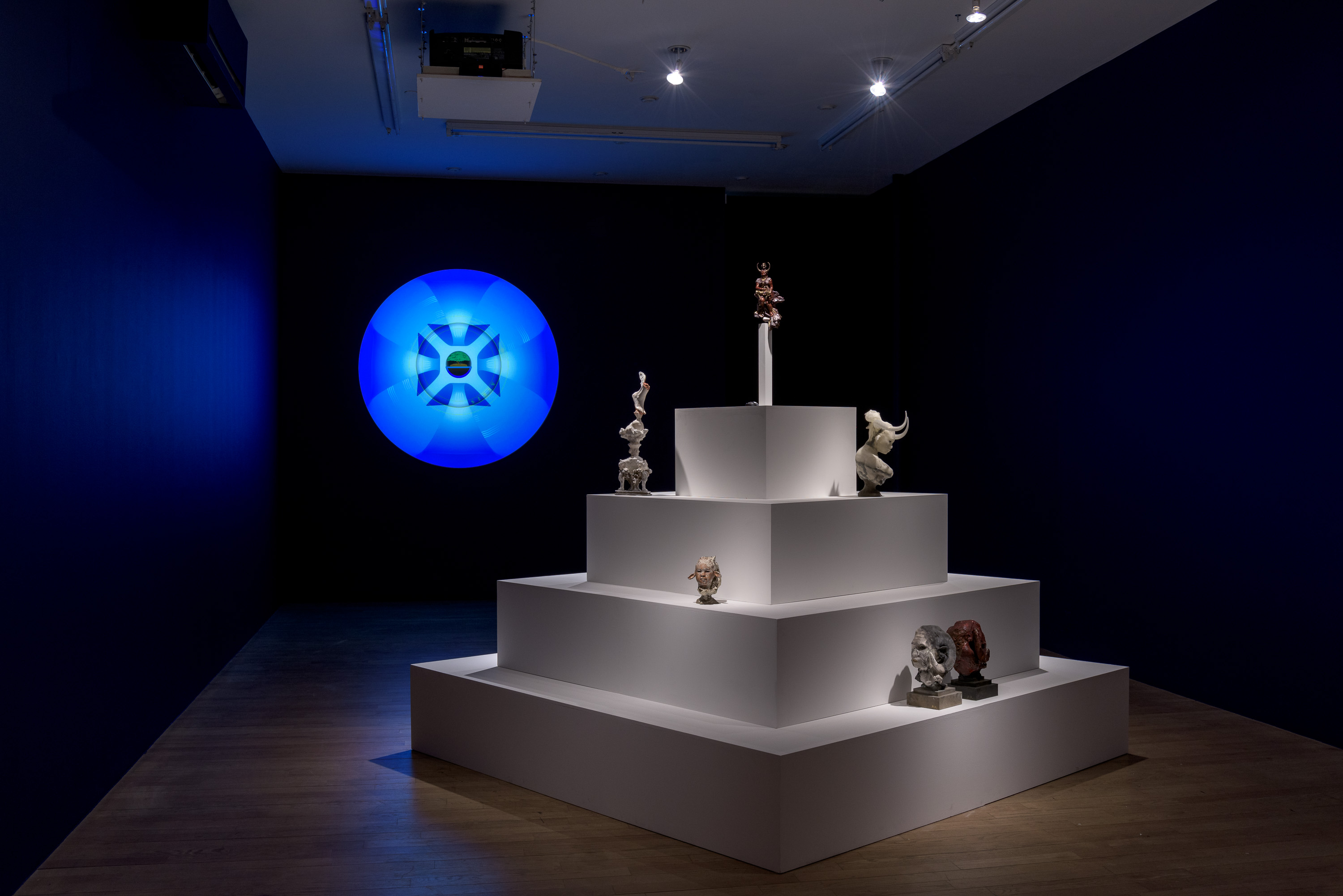 Auriea's Year Zero at bitforms, New York: a stepped white ziggurat of sculptures — horned bust, small heads, a crowned figure on a column — beside a glowing blue mandala projection