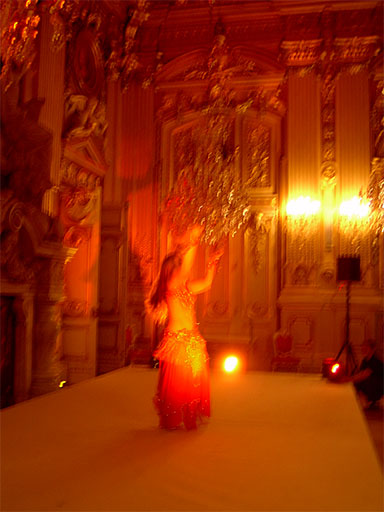 Blurred photograph in amber light: a bellydancer in a sequined costume performs on a white runway in an ornate gilded hall